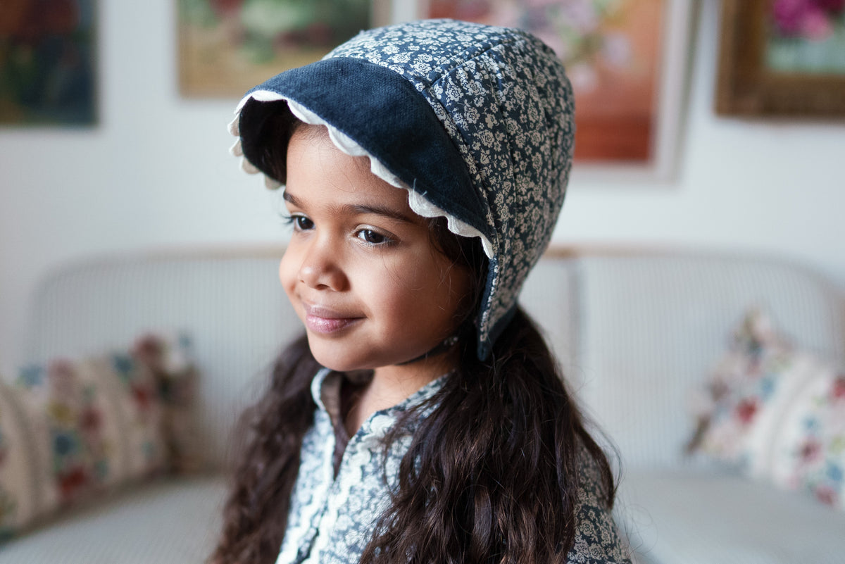 Girl smiling wearing pretty floral bonnet with cord and scalloped details with matching coat in a vintage inspired setting.