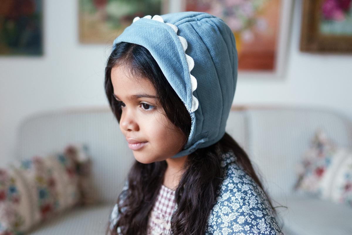 Girl smiling wearing vintage inspired bonnet in eggshell blue with rim pushed back with scallop detailing, showing vintage-inspired outfit.