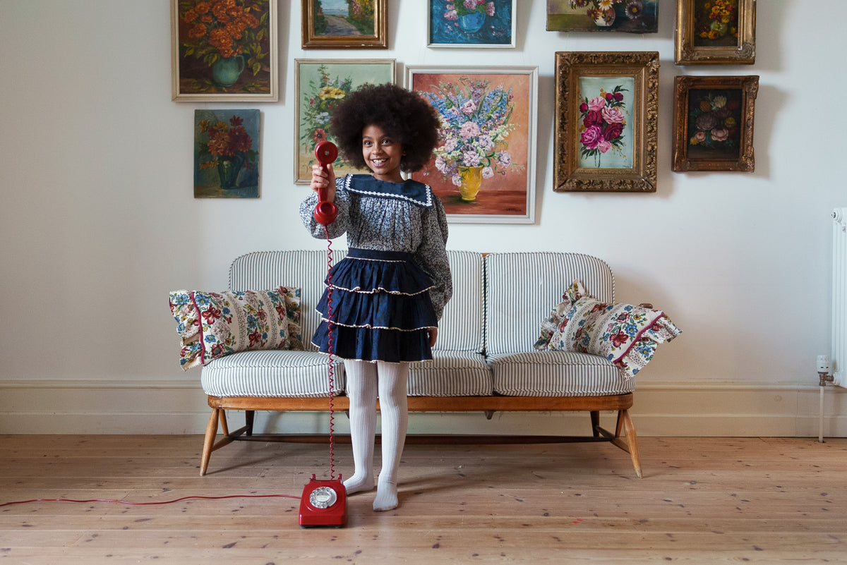 Young girl smiling holding up classic phone wearing navy floral vintage-inspired blouse with velvet sailor collar and velvet rara skirt made form organic  fibers in a rustic setting showcasing sustainable values.
