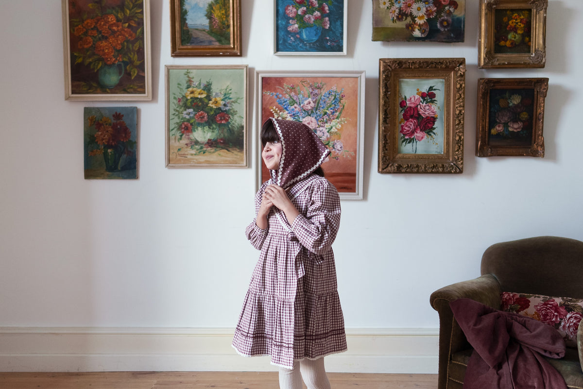 Young girl wearing plum gingham wrap dress and cream tights, smiling with plum floral scarf draped over head, in front of classic flower paintings in a vintage setting.