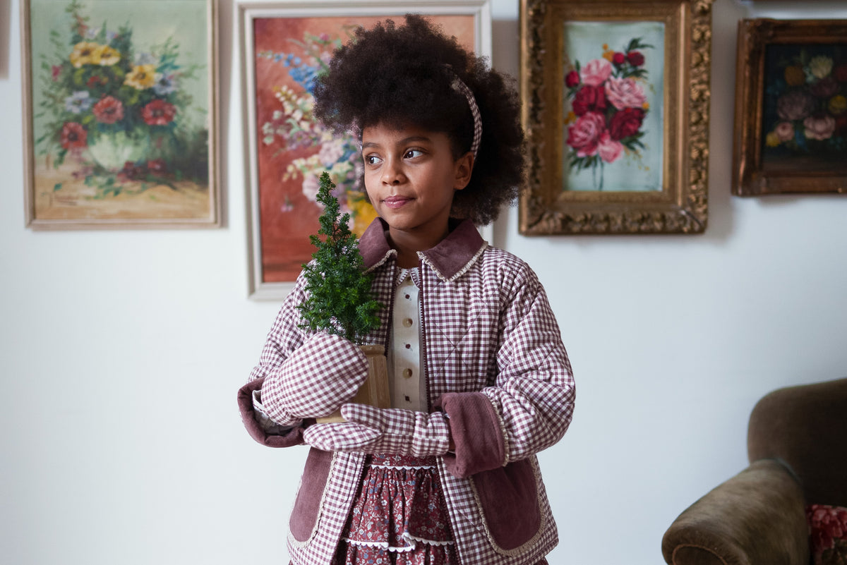 Child smiling holding small fir tree wearing plum organic cotton gingham coat with contrasting pockets and collar and matching mittens, for sustainable children's clothing.