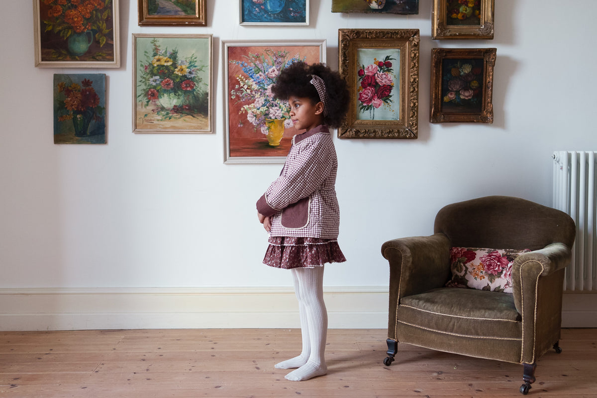 Child's profile wearing plum gingham coat with pockets and floral skirt with cream tights, stood on rustic wooden floor with flower paintings on wall behind, showcasing a vintage inspired setting.