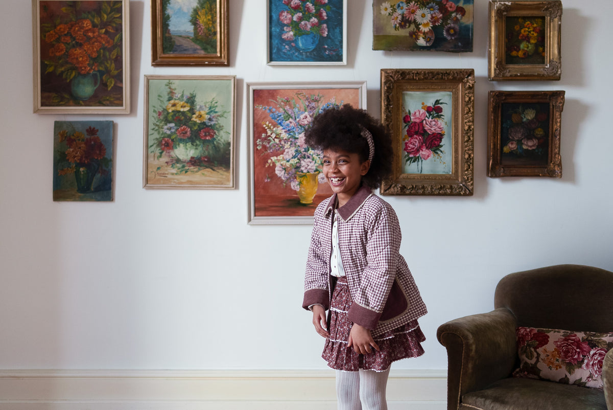 Young girl smiling wearing plum gingham winter coat and red floral rara skirt all made from organic materials showcasing sustainable and ethical values.