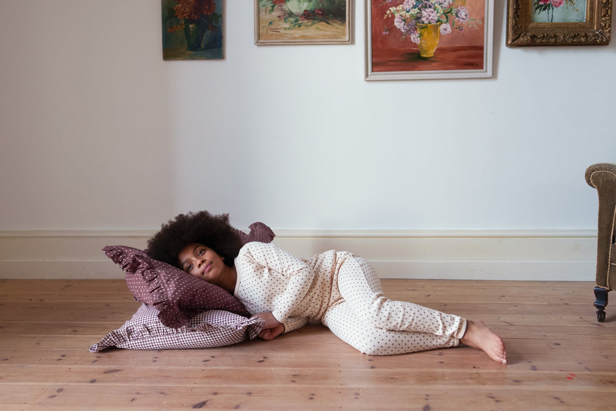 Girl laying on rustic wooden floor with red floral and gingham cushions, wearing cream and red ditsy floral pyjama set made from organic cotton, showing sustainable values in a vintage inspired setting.