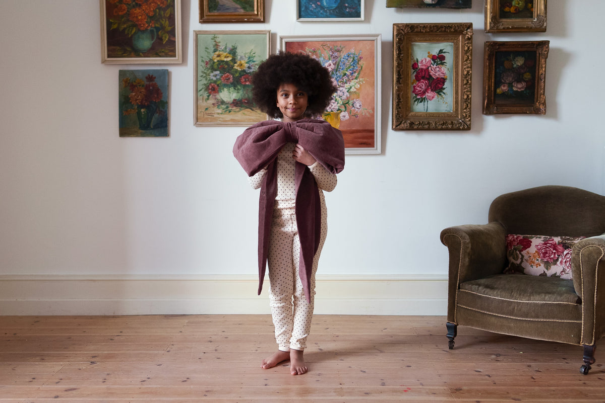 Girl stood on wooden floor holding large velvet red bow, wearing organic cotton pyjama set, stood in front of classic flower paintings on wall in vintage frames.