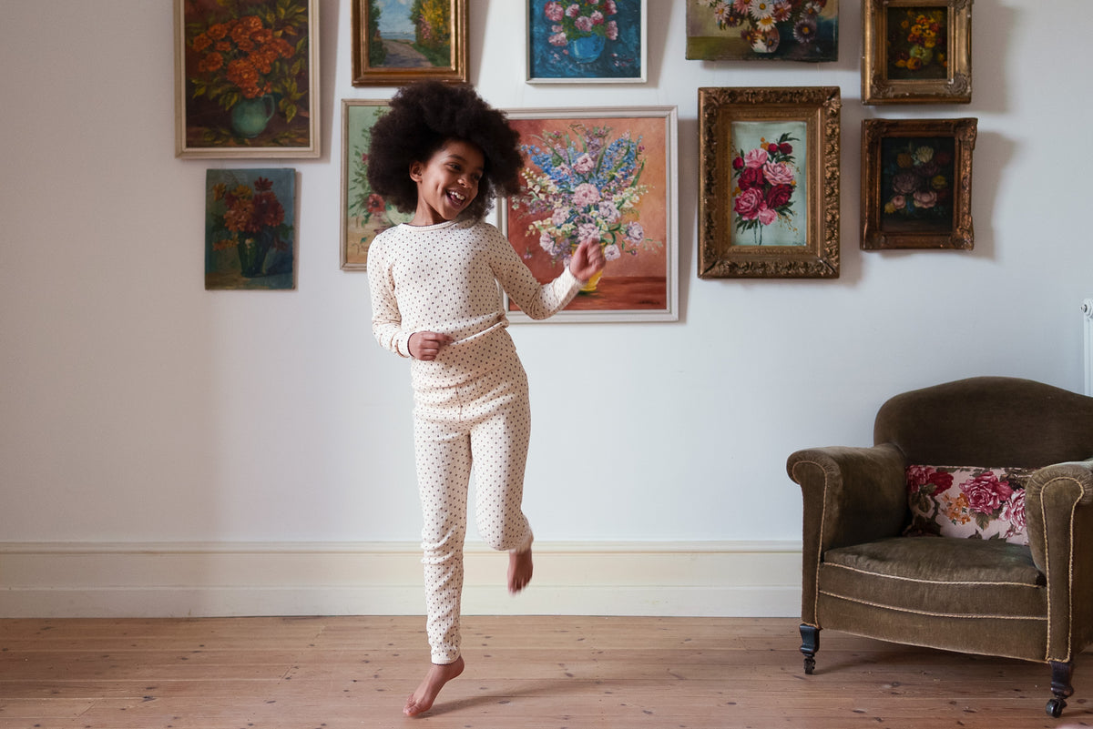 Young girl dancing wearing cream and red and ditsy floral print pyjama set, on rustic wooden floor with classic flower paintings on the wall, showing a vintage-inspired setting. 