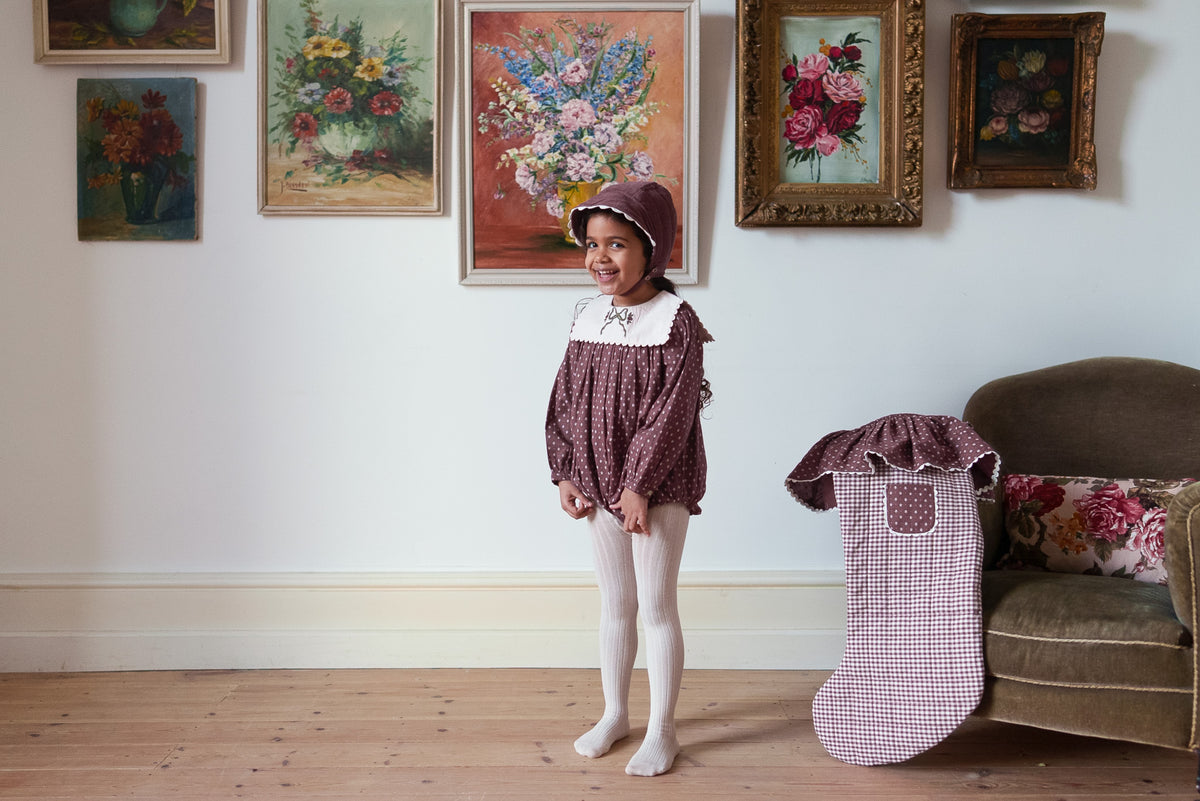 Girl smiling wearing sugarplum bonnet and matching romper made from organic cotton paired with cream tights showing a vintage-inspired outfit on rustic wooden floor with floral paintings behind, showing a classic setting and sustainable values.