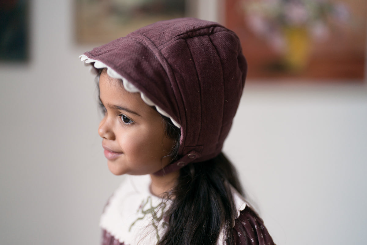 Girl smiling wearing plum velvet vintage-inspired bonnet with white scallop edging and matching romper, all made from organic cotton showing sustainable values.