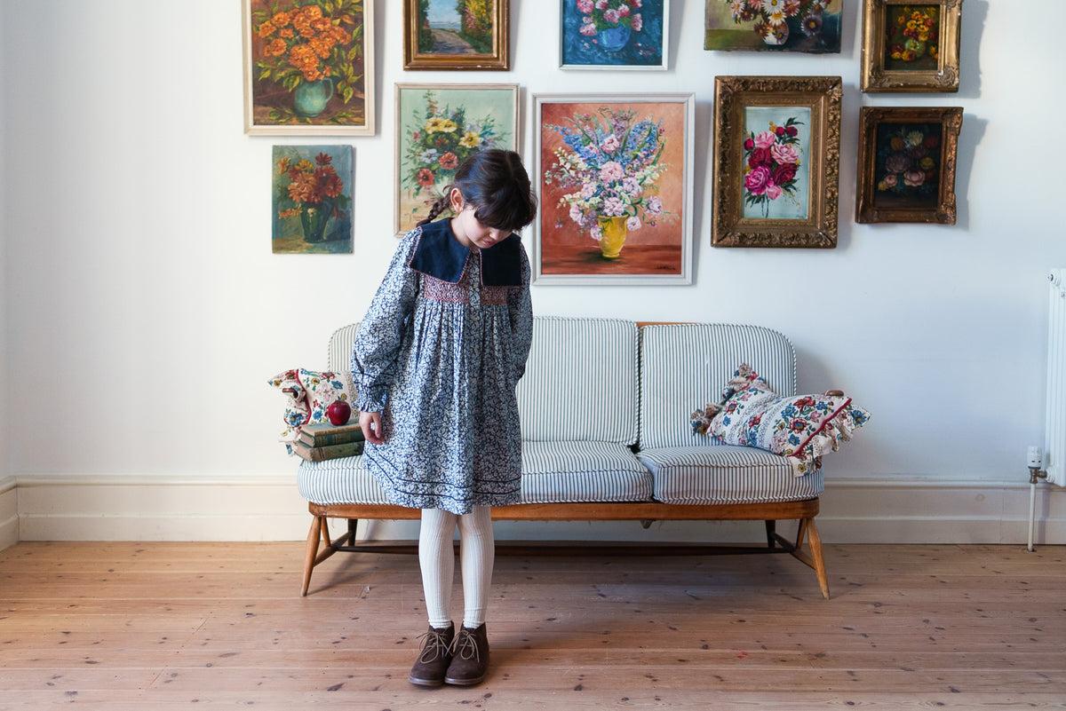 Young girl wearing vintage-inspired floral dress with long sleeves and velvet sailor collar paired with cream tights, stood on rustic wooden floor with vintage inspired decor showing sustainable children's clothing.