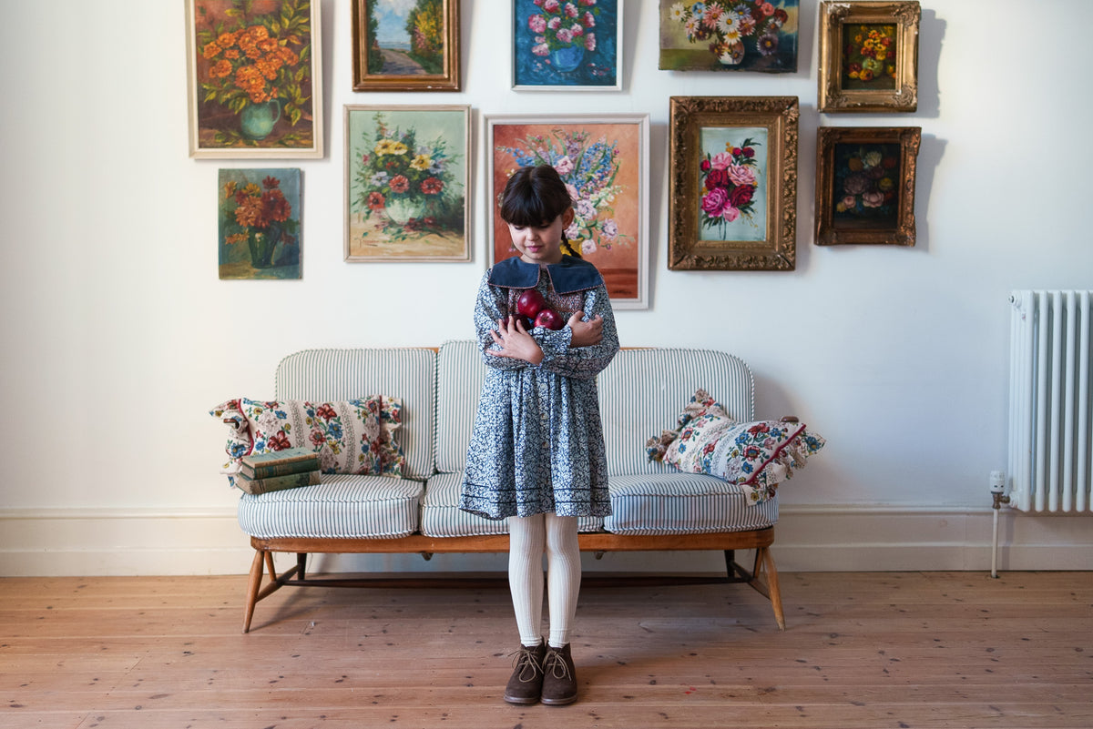 Young girl with armful of apples wearing vintage inspired floral dress made from organic cotton with velvet sailor collar and cream tights stood against rustic sofa with floral paintings on wall, showing sustainable kids clothing.