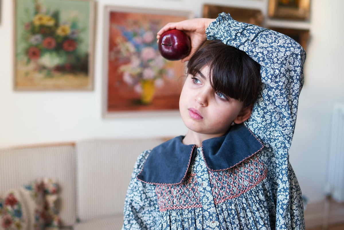 Young girl holding apple with arm over her head in vintage-inspired floral dress with contrasting pink smocking and velvet sailor collar design, made from organic cotton for ethical and sustainable children's clothes.