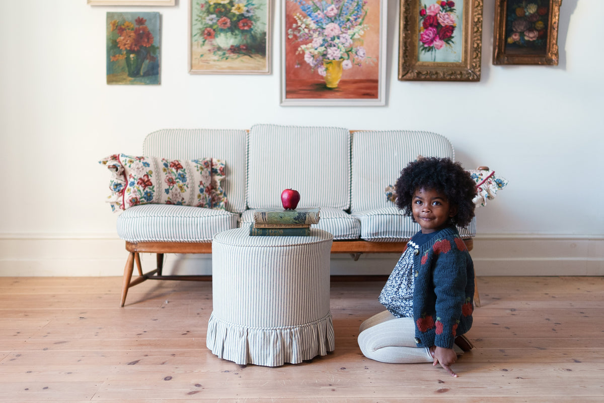 Child sat on floor smiling at camera in navy floral romper with handknit apple patterned cardigan, made from natural fibers surrounded with vintage decor, showcasing sustainable children's clothing.