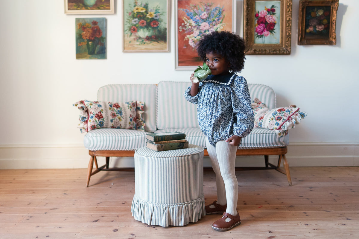 Child wearing navy and white floral romper with velvet sailor collar and contrasting ric rac trim, stood next to vintage inspired striped furniture showing sustainable clothing.