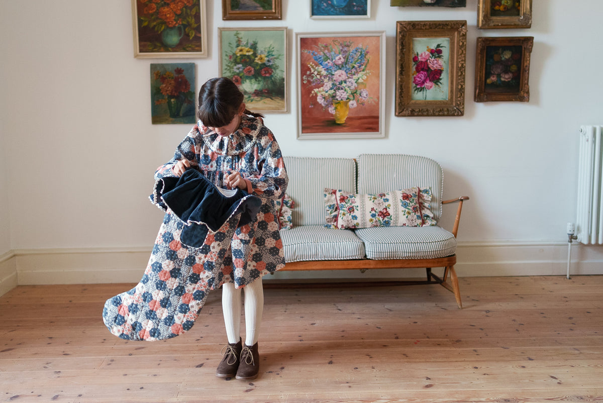 A girl stands in a room with wooden floors and a sofa, holding up a long, patterned fabric item that appears to be a scarf or wrap. The fabric is a patchwork priny