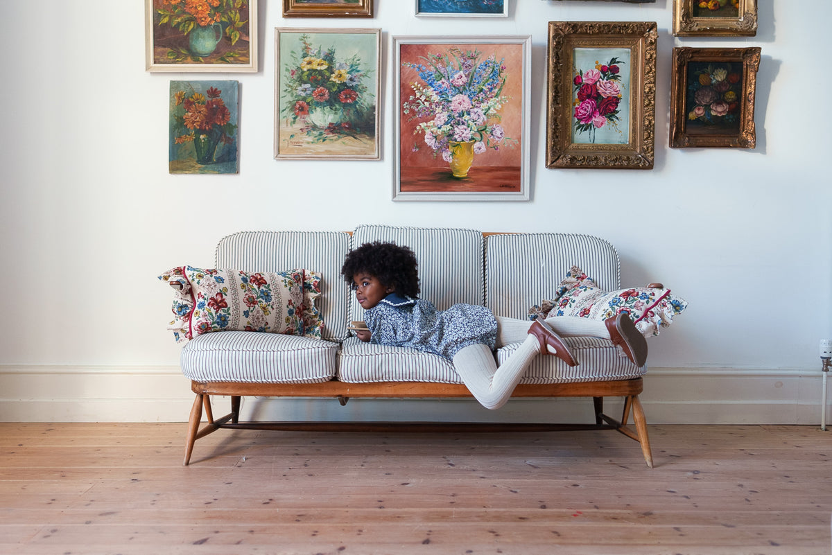 Young child wearing navy and white vintage inspired romper with floral pattern and sailor collar made from organic cotton, lounging on sofa in front of vintage floral paintings, showing sustainable children's clothing.