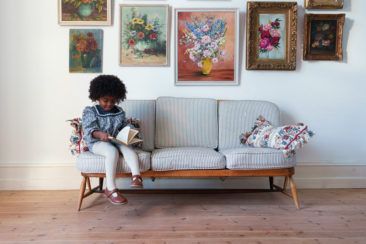 Young child reading book wearing navy and white floral vintage-inspired romper with velvet sailor collar, sat on striped sofa in front of wall with floral paintings.