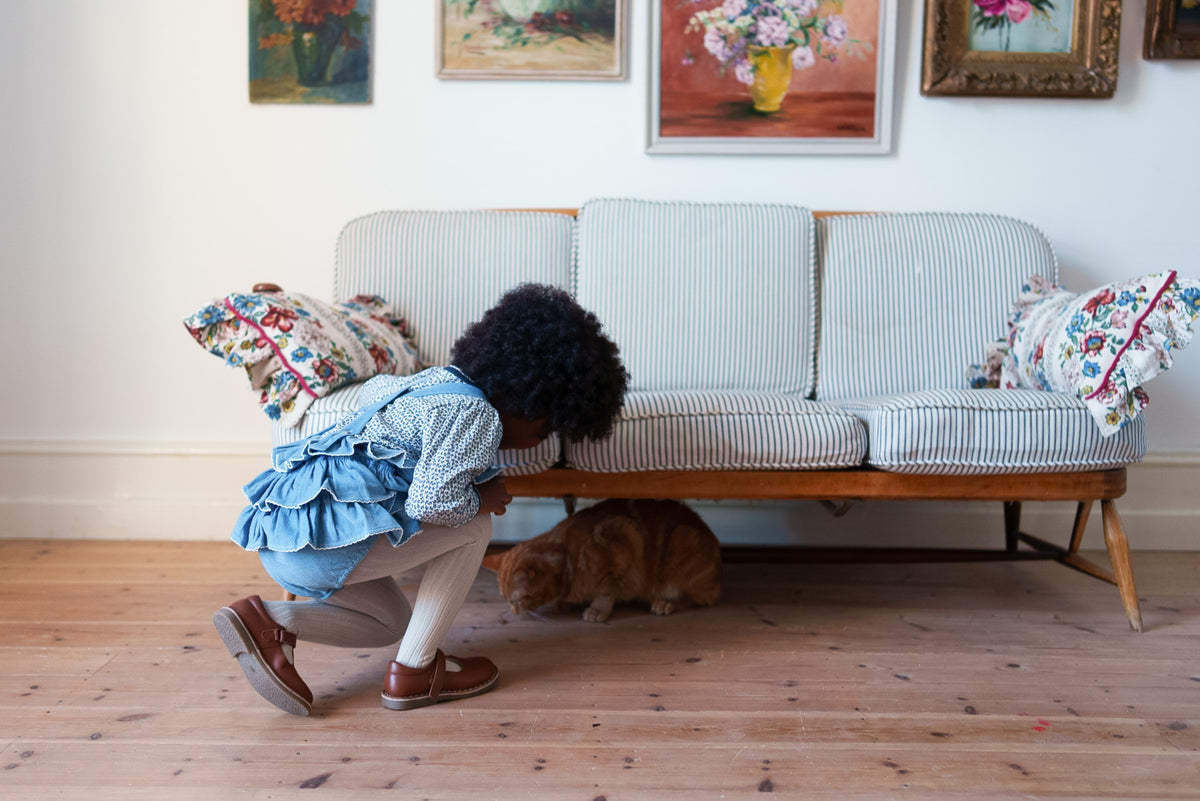 Child wearing organic cotton floral t-shirt layered underneath blue velvet frilly romper, crouching on rustic wooden floor looking beneath a sofa at a ginger haired cat.