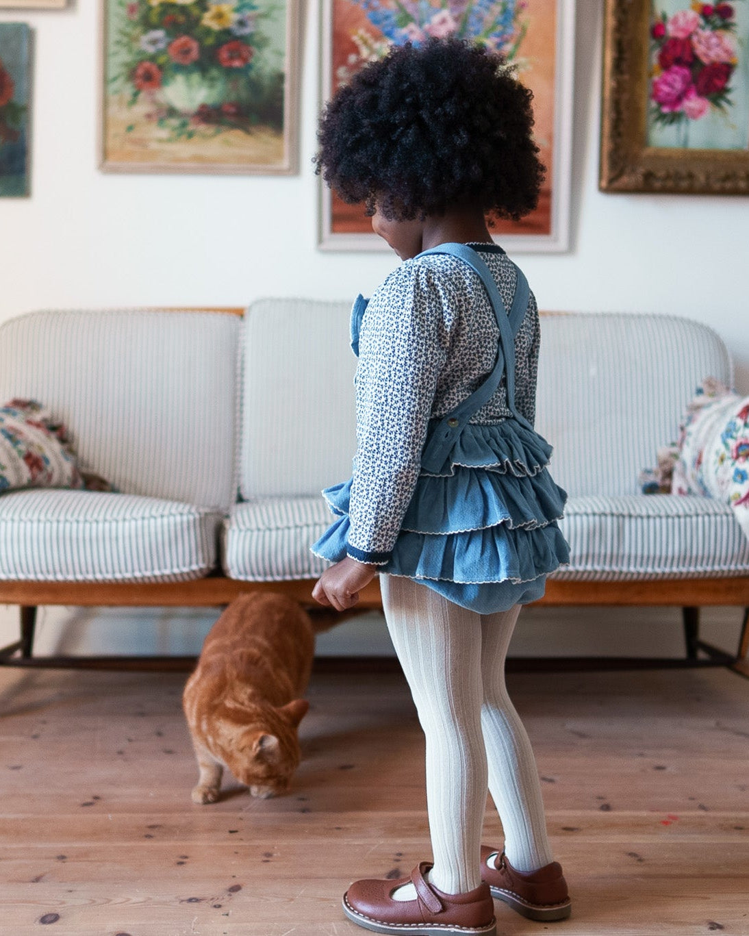 Child wearing a vintage style velvet romper with white tights and brown shoes beside a dog in a cosy sitting room with floral artwork