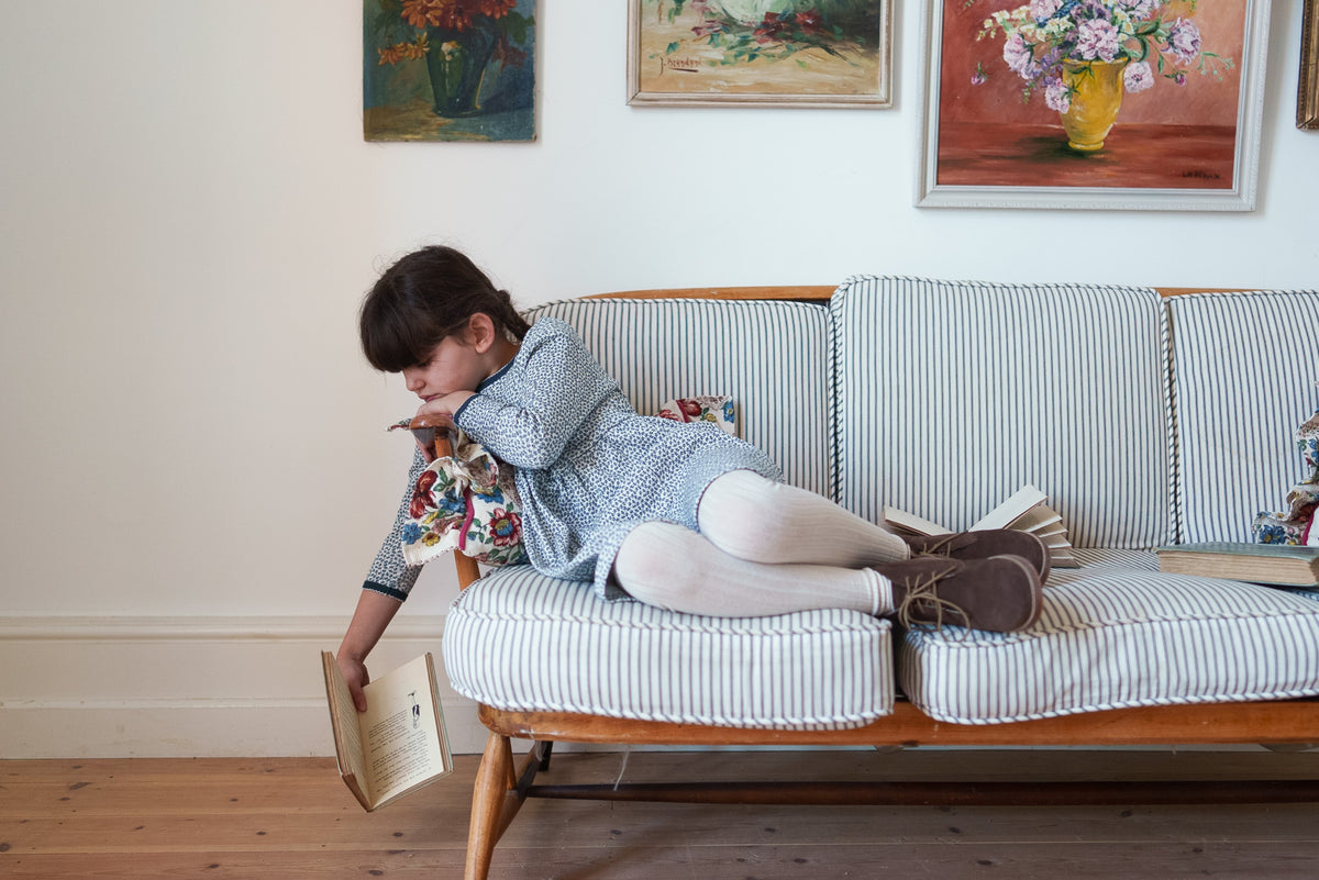 Young girl wearing black and white floral organic cotton dress paired with cream tights and brown boots, laying across striped sofa in a vintage-inspired setting, representing sustainable values.