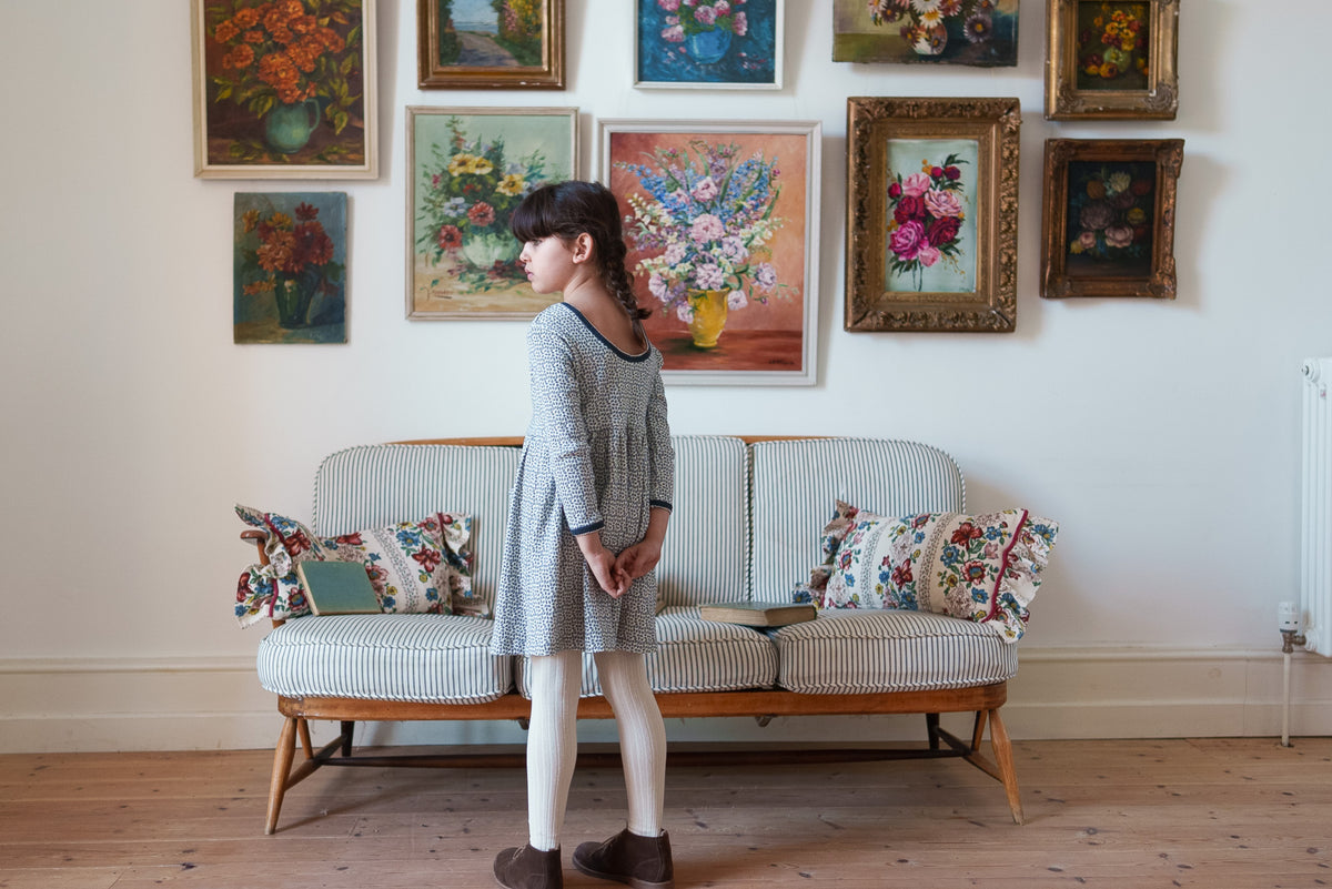 Young girl wearing low backed soft cotton jersey floral dress with long sleeves and cream tights, stood on rustic wooden floor in front of striped sofa with flower paintings on the wall.