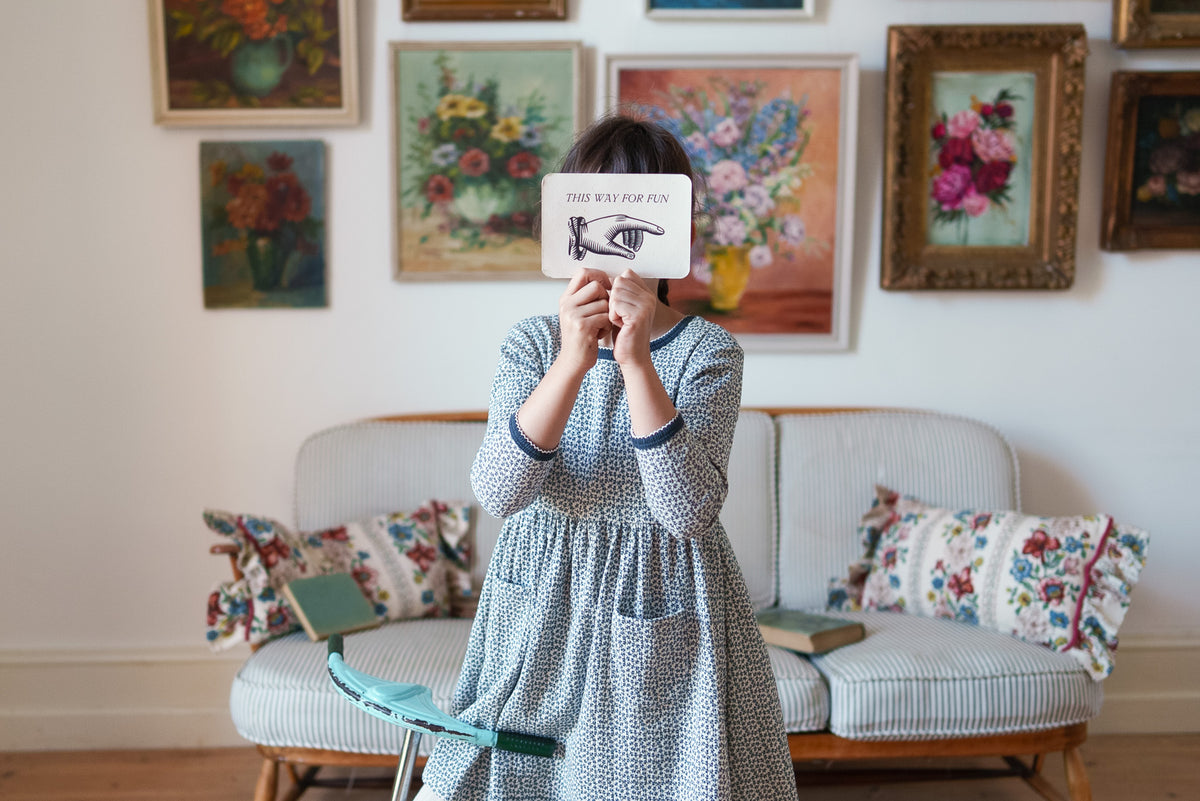 Young girl wearing black and white floral pointelle dress made from organic cotton, holding card in front of her face, stood in front of sofa in a vintage setting.