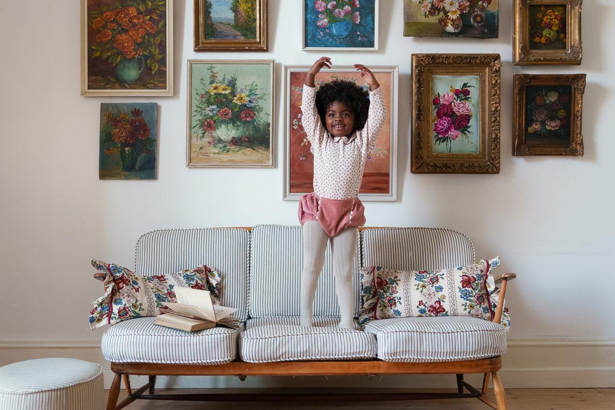 Young girl stood on sofa with arms raised wearing soft pink cord bloomers and cream tights with matching top all made from organic cotton, in front of classic flower paintings showing a vintage inspired setting for sustainable children's clothes.