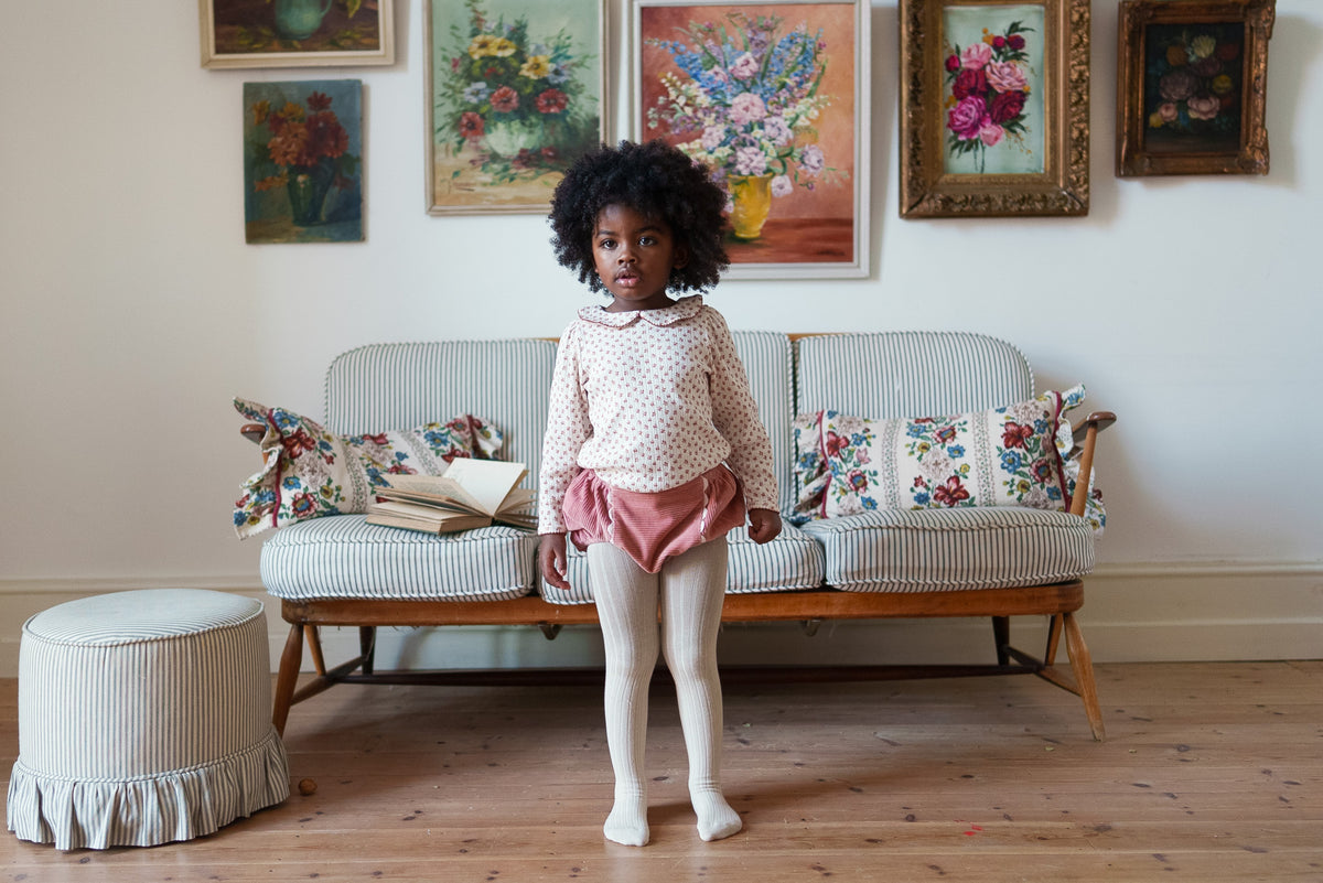 Young girl stood wearing soft pink cord bloomers with cream floral top and cream tights, made from organic cotton showing sustainable values in front of vintage inspired decor.