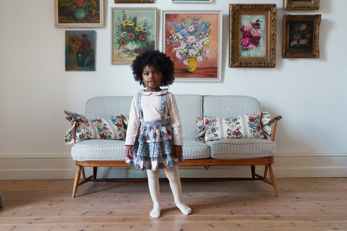 Young girl wearing patchwork floral rara skirt with cotton top underneath and cream tights, stood on rustic wooden floor in front of vintage-inspired decor.