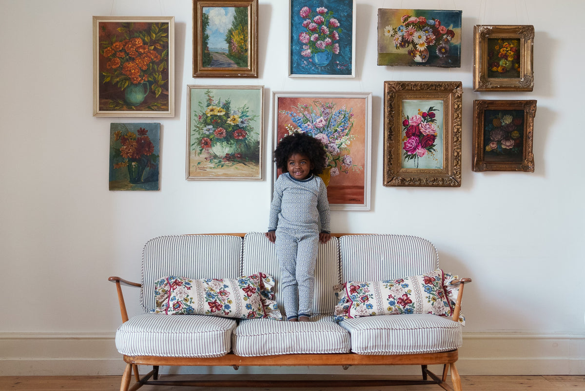 Young child smiling wearing black and white floral pointelle pyjama set, stood on striped sofa against a white wall with vintage-inspired flower paintings.