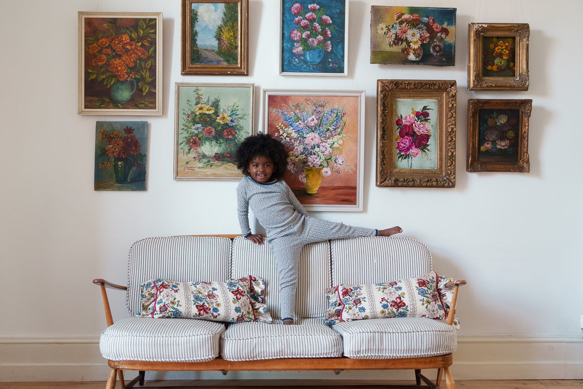 Young child sitting on back of striped sofa wearing organic cotton black and white floral pyjama set with classic flower paintings in the background.