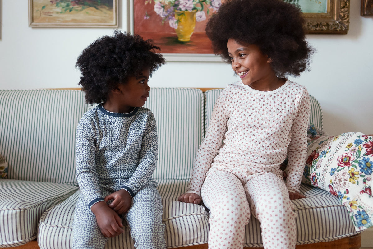Two children smiling at each other wearing floral organic cotton pyjama sets, sat on striped sofa with flower paintings in background.
