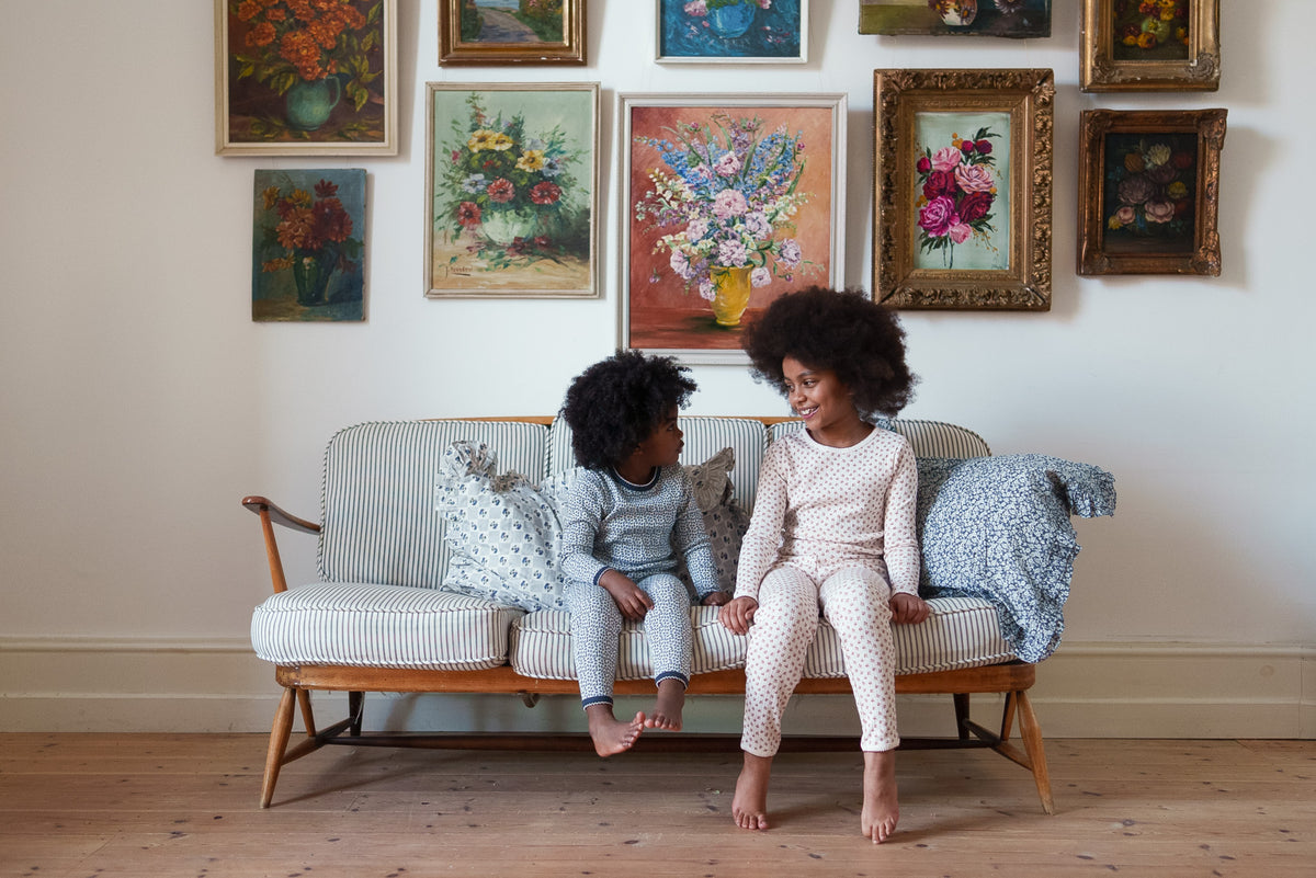 Two children wearing cotton floral pyjama sets smiling at each other while sat on striped sofa and classic flower paintings on wall behind them, displaying a vintage-inspired setting.