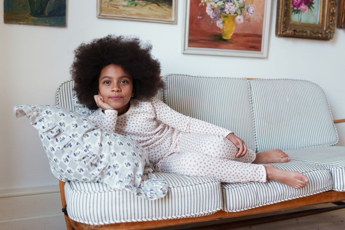 Young girl looking at camera sat on striped sofa, wearing cream and red festive pyjamas made from organic cotton jersey, showing sustainable values for children's clothes.