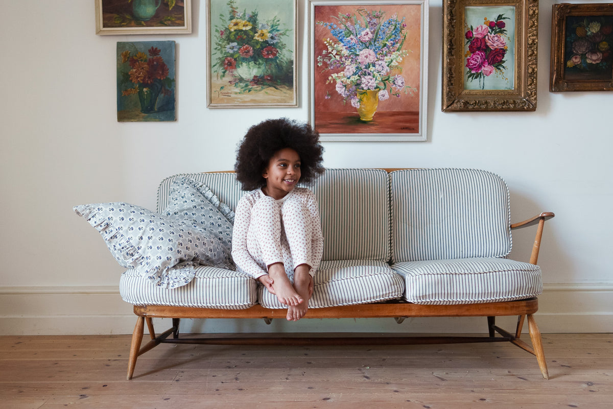 Girl sat on sofa with knees up wearing cream and red floral organic cotton pyjama set, sat on striped sofa with pretty flower paintings on the wall behind.