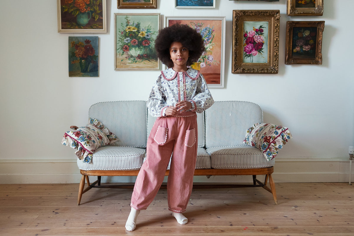 Young girl wearing loose pink cord trousers with pockets and white floral blouse with matching pink trim, made from organic cotton for sustainable children's clothes.
