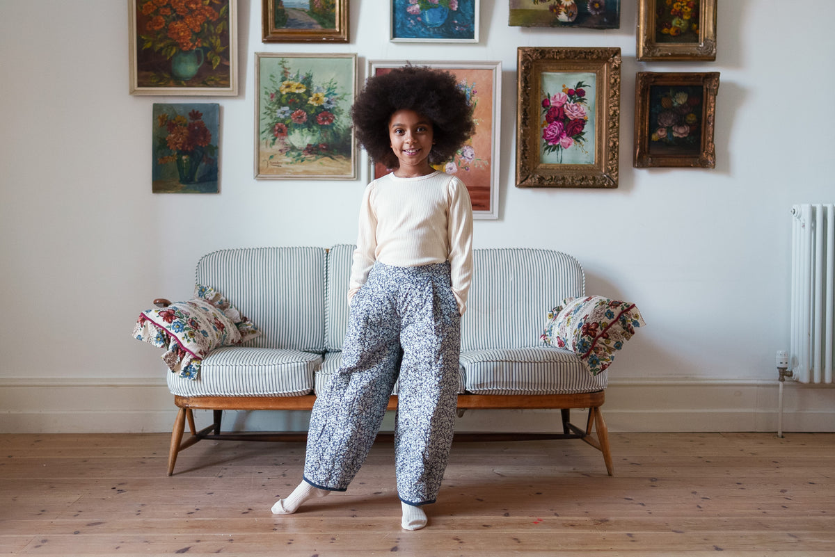 Girl wearing cream organic cotton long sleeved top with quilted floral trousers, stood in front of striped sofa with floral decorations.
