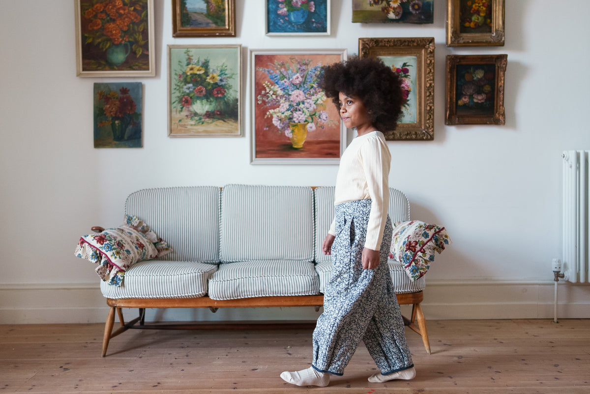 Young girl wearing quilted black and white floral trousers with cream soft organic long sleeved top and matching socks, walking past striped sofa with classic flower paintings on wall in a vintage setting.