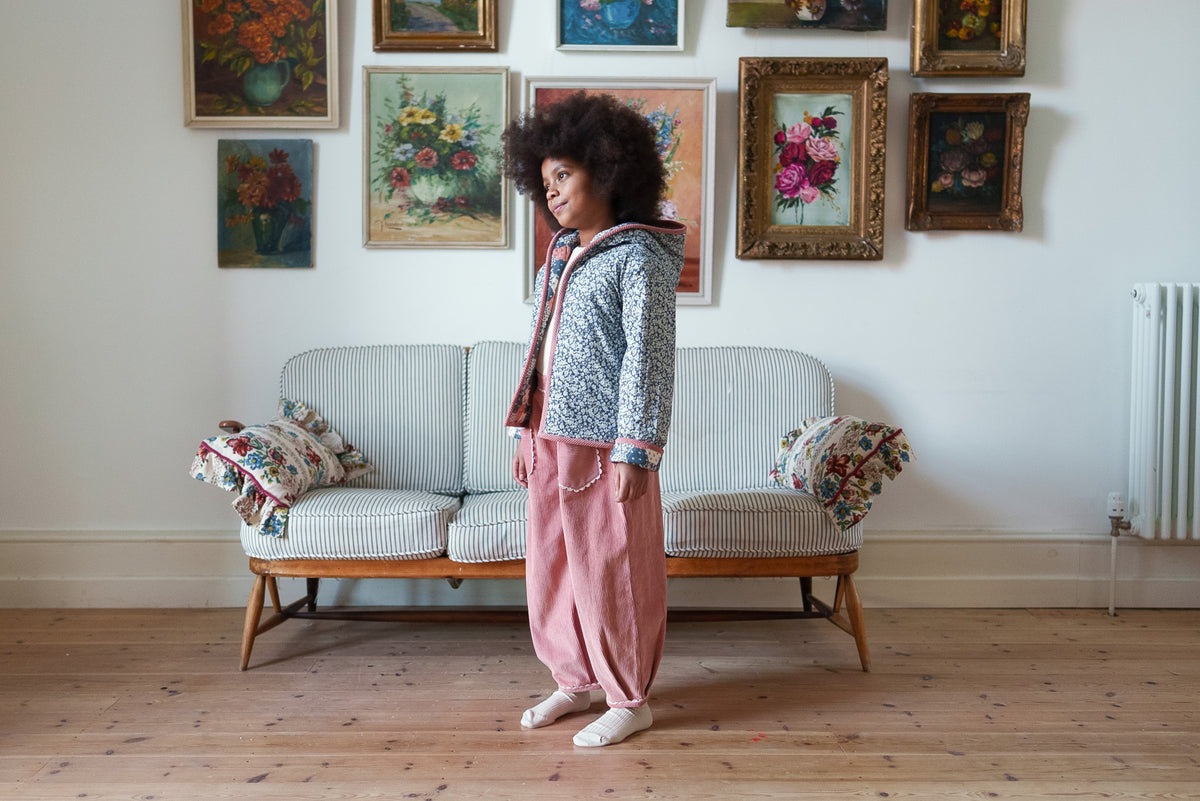 Young girl wearing loose pink cord trousers and floral patchwork coat  stood against rustic striped sofa with flower paintings on wall behind, showing a vintage inspired children's clothing brand.