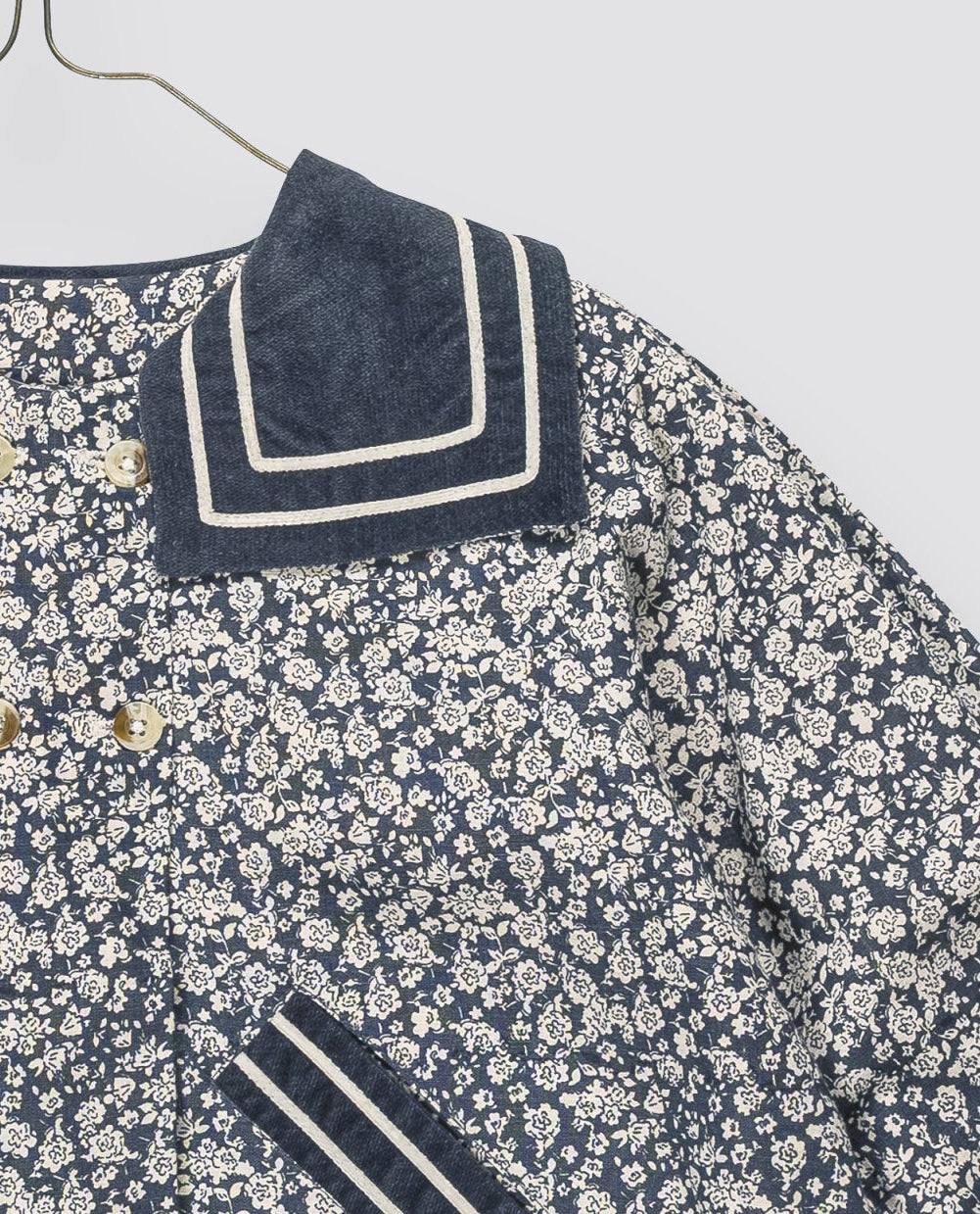 A children's long-sleeve jacket with a navy blue and cream floral print, featuring solid navy blue collar and cuff trim with cream stripes, displayed on a metal hanger against a light gray background.