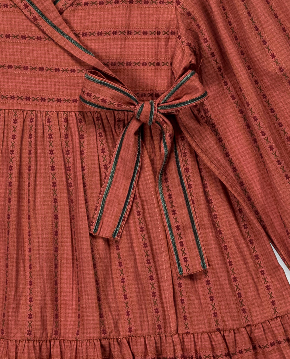 A terracotta-colored long-sleeve wrap dress with thin black vertical stripes, featuring a tiered skirt and a matching fabric bow at the waist, hanging on a metal hanger against a light gray background.