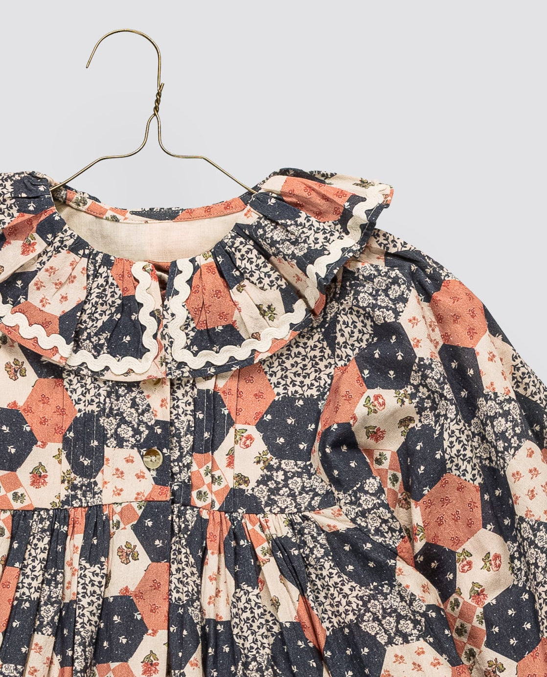 A long-sleeve baby dress with a ruffled collar, featuring an all-over patchwork floral and geometric print in navy, rust orange, and cream. The dress is shown hanging on a simple metal hanger against a solid light gray background.