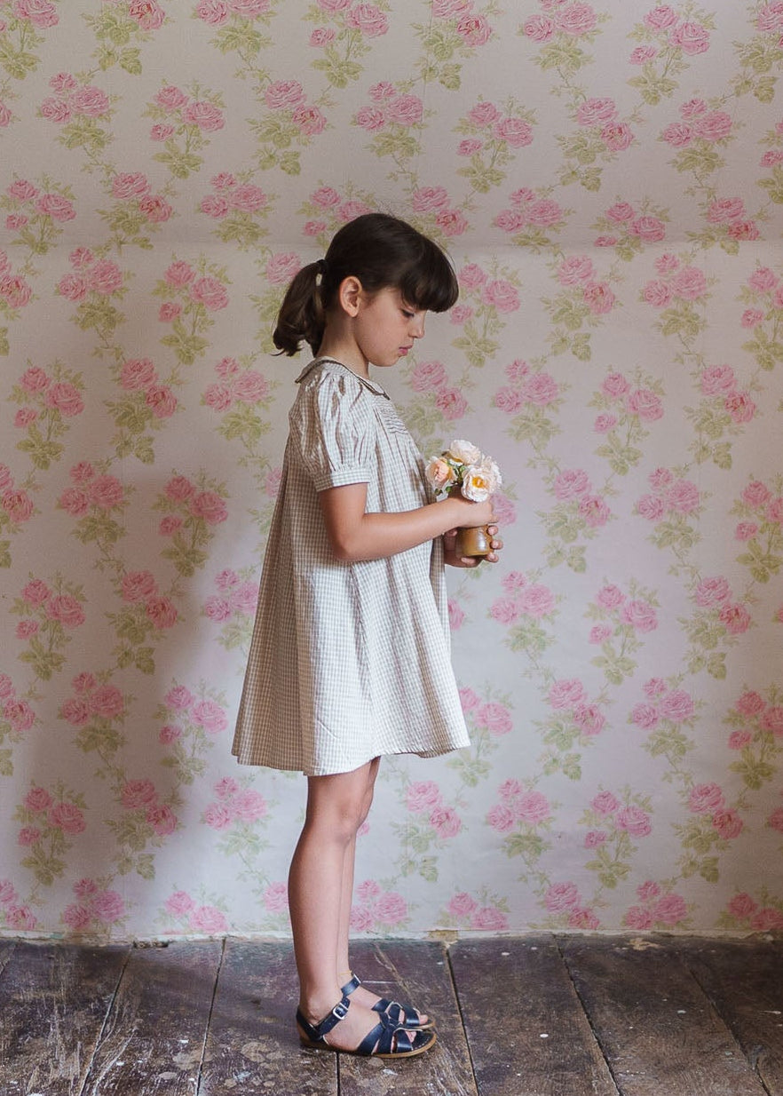 Young girl wearing a vintage-style cotton blend checkered dress holding a small bouquet, standing against a floral wallpaper background in a classic, natural setting.