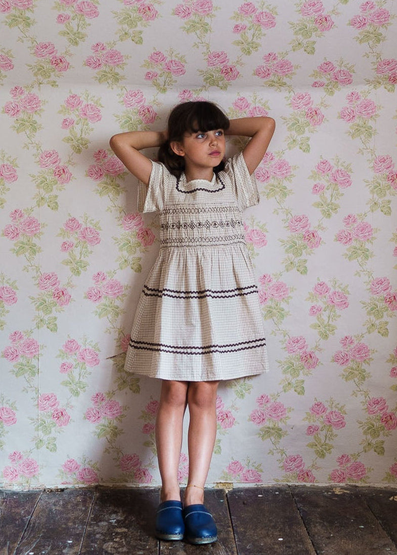 Young girl wearing a vintage style smocked dress in a yellow check pattern made from a cotton blend, standing against a floral wallpaper background, showcasing sustainable children's clothing.