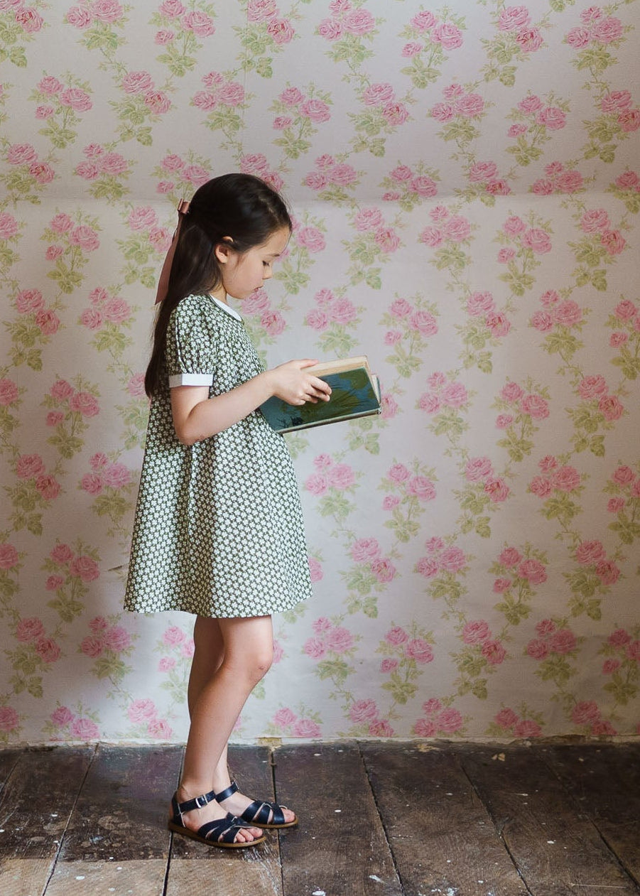 Young girl wearing a green floral cotton blend dress with a vintage style, reading a book against a backdrop of pink floral wallpaper, embodying natural and sustainable kids clothing.