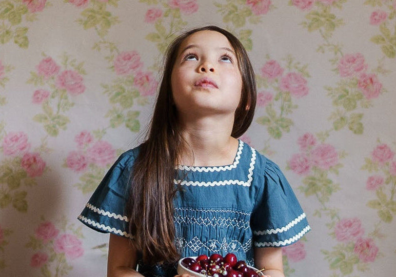 Young girl wearing a vintage style organic cotton blue dress with smocked detailing, sitting on a wooden box holding a bowl of cherries against floral wallpaper.