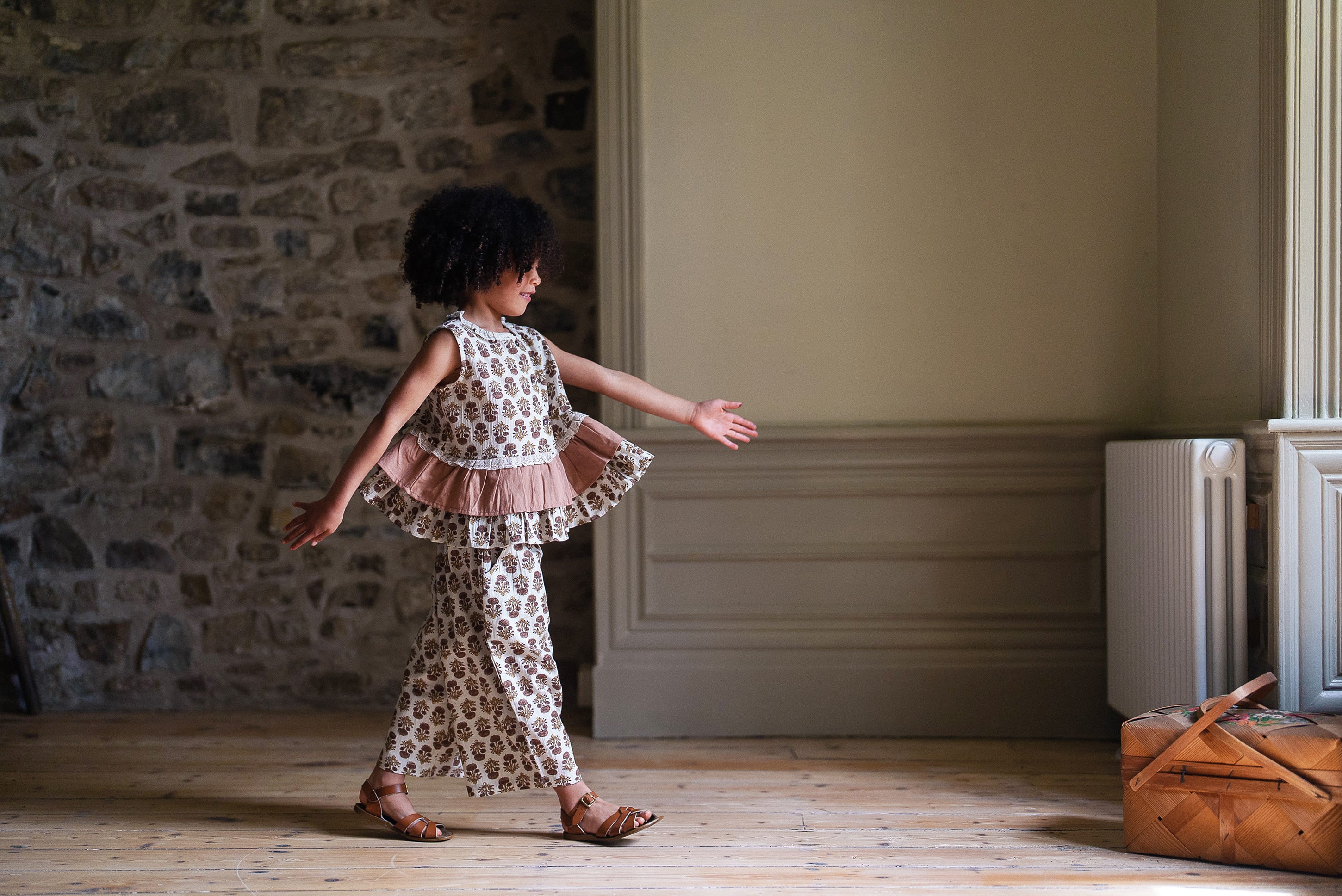 Child wearing vintage style organic cotton clothing with floral patterns and tea rose accents, walking indoors on wooden floor in soft natural light.