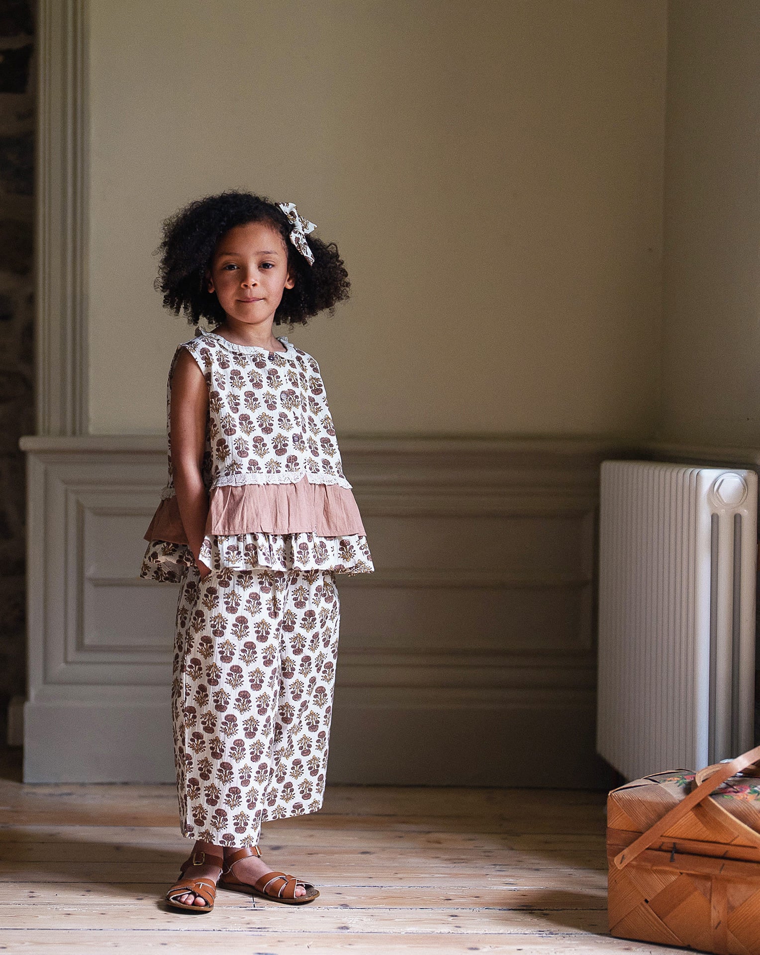 Young girl wearing vintage style organic cotton blouse and matching wide-leg pants with a floral pattern and tea rose accents, standing indoors in natural light.