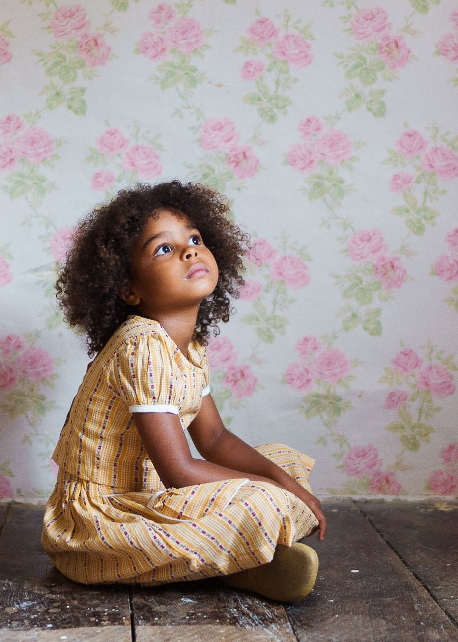 Young girl with curly hair sitting on a wooden floor wearing a yellow floral organic cotton top and skirt, vintage-inspired children’s clothing in a soft natural setting.