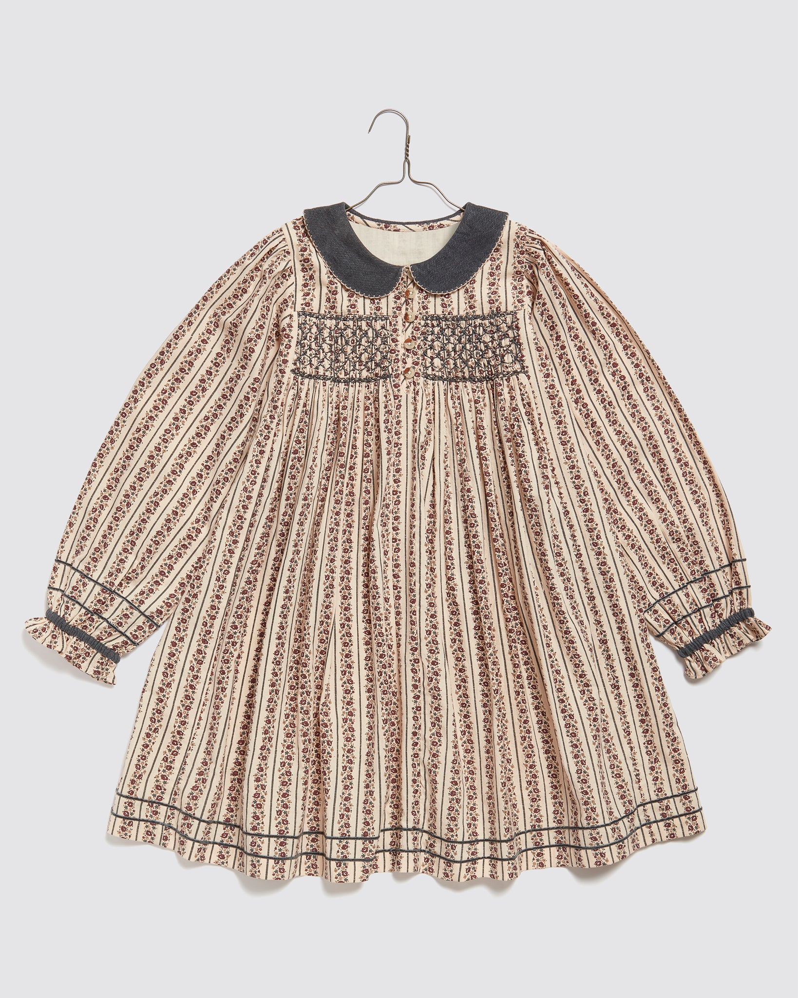 Smocked vintage-style girls dress made from organic cotton with a floral and stripe pattern, featuring a dark Peter Pan collar and long puffed sleeves.
