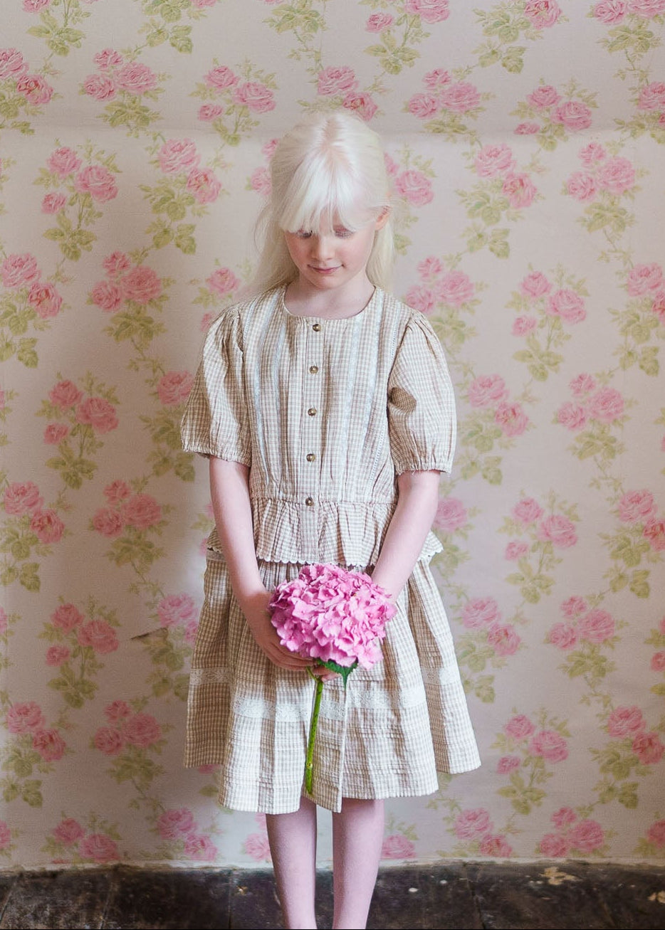 Young girl wearing a cream checkered organic cotton skirt and matching top, holding a pink flower against a vintage floral wallpaper backdrop, embodying natural, sustainable children's clothing.